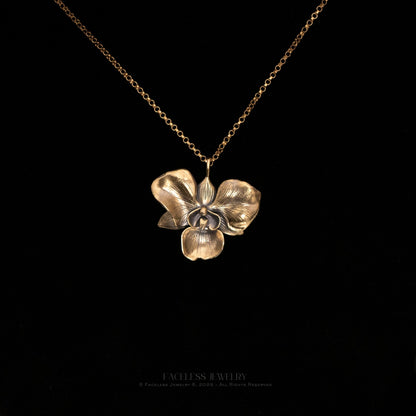 The Golden Orchid pendant by Faceless Jewelry displayed against a black background, featuring a 14Kt gold orchid flower with finely detailed petals and a warm, polished finish, hanging from a delicate gold rolo chain.