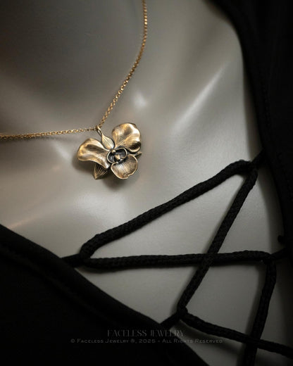 The Golden Orchid pendant by Faceless Jewelry displayed on a mannequin wearing a black lace-up top, featuring a 14Kt gold orchid flower with soft, curved petals and detailed texture hanging from a fine gold chain near the collarbone.