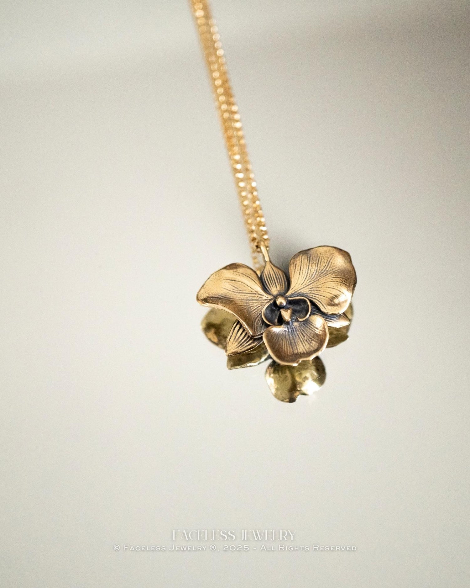 The Golden Orchid pendant by Faceless Jewelry resting on a mirror, featuring a 14Kt gold orchid flower with detailed petals and a brushed finish, its reflection softly visible beneath the gold chain.